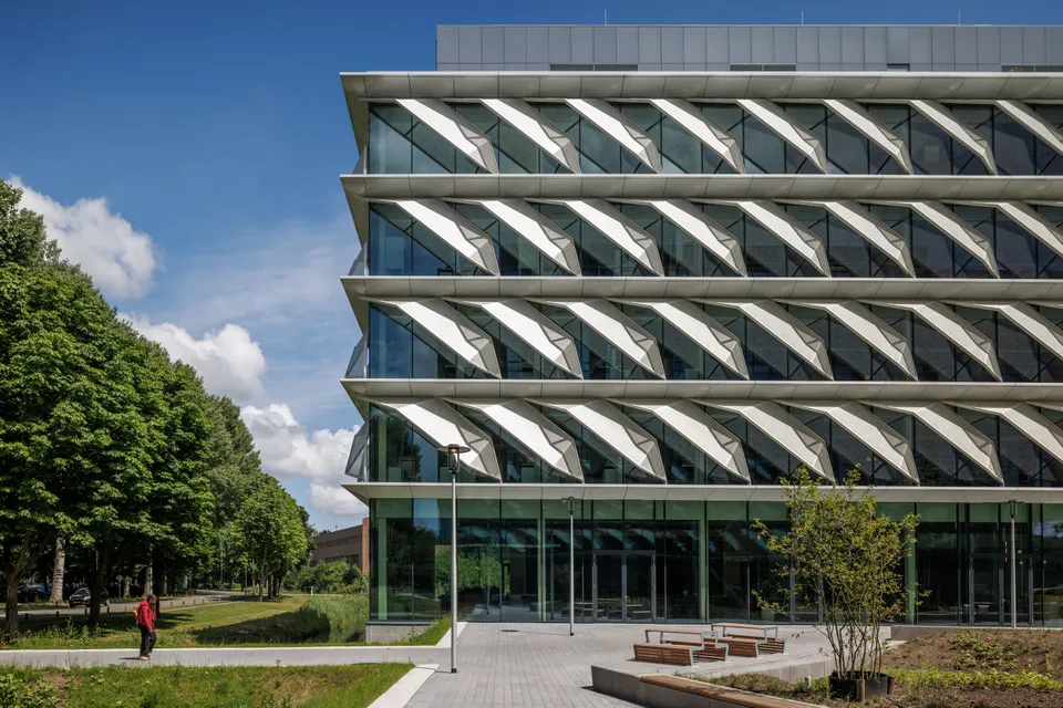 Modern glass building with angled windows and greenery. A person in red walks on a path nearby under a blue sky with clouds.