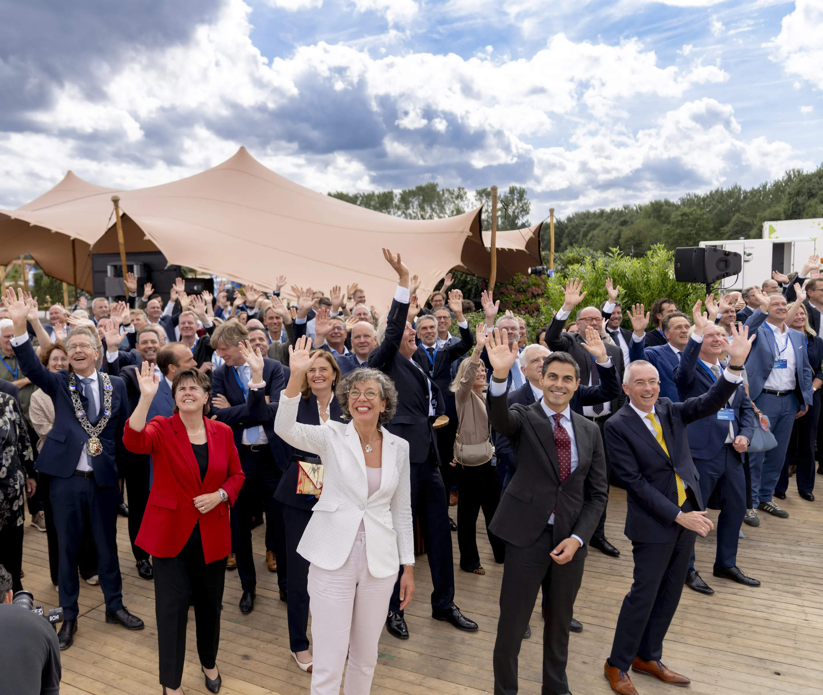 A large group of people, wearing formal attire, smiling and waving outdoors under a cloudy sky, standing on a wooden deck near tented structures.