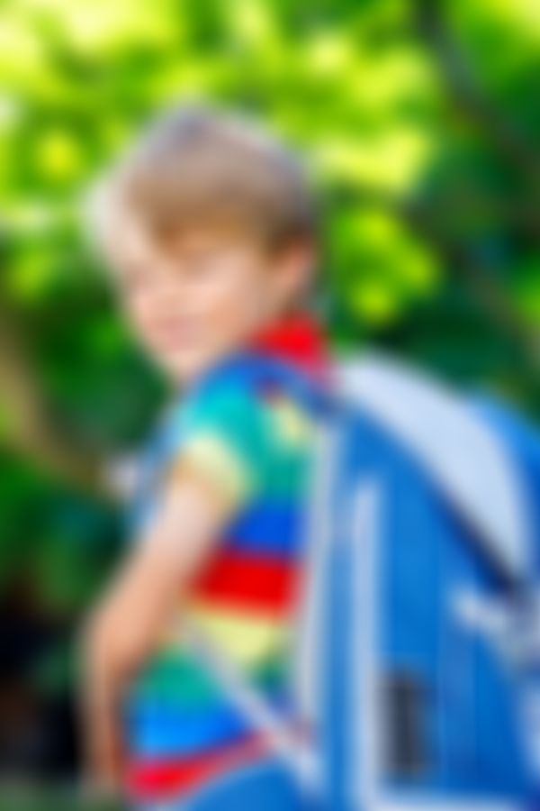 Junge mit buntem T-Shirt und blauer Schultasche lächelt in die Kamera, Hintergrund mit grün verschwommenem Laub.