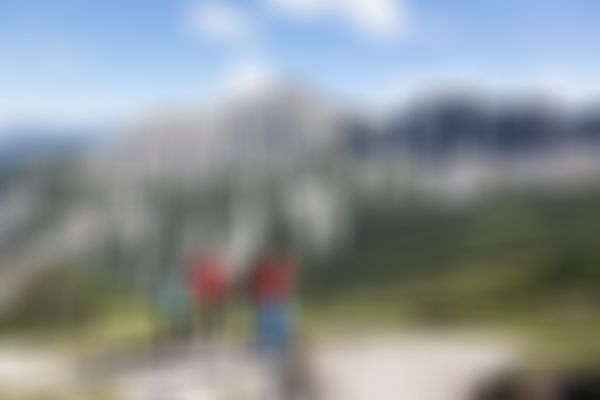 Drei Wanderer in farbenfroher Kleidung wandern auf einem felsigen Pfad mit schroffen Bergen und einem grünen Tal im Hintergrund unter einem klaren blauen Himmel.