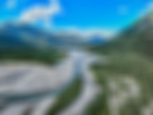 Luftaufnahme des sich schlangelnden Lechs durch das saftig grüne Lechtal, eingerahmt von dicht bewaldeten Bergketten unter einem strahlend blauen Himmel mit leichten Quellwolken.