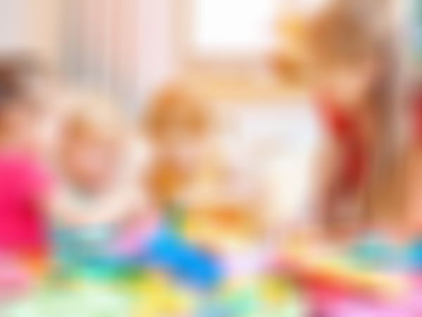 Drei kleine Kinder spielen gemeinsam mit bunten Holzbausteinen, während eine Erzieherin in rotem Kleid sie lächelnd begleitet.