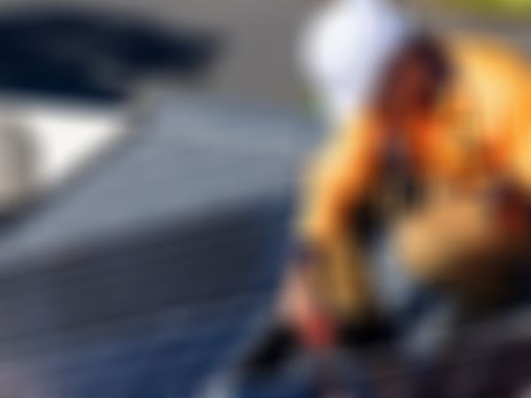 Ein Arbeiter in orangener Sicherheitsjacke und Schutzhelm montiert Photovoltaikmodule auf einem Dach mit einem roten Akkuschrauber.