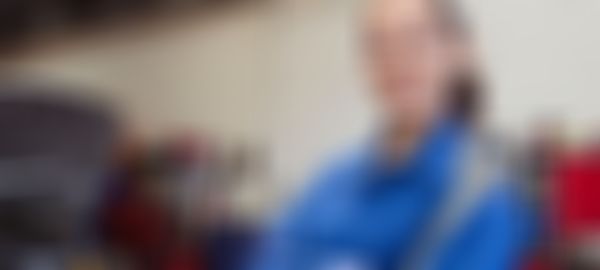 Female mechanic in blue coveralls holds a large wrench, standing confidently in a garage with a car hood open and tools in the background.