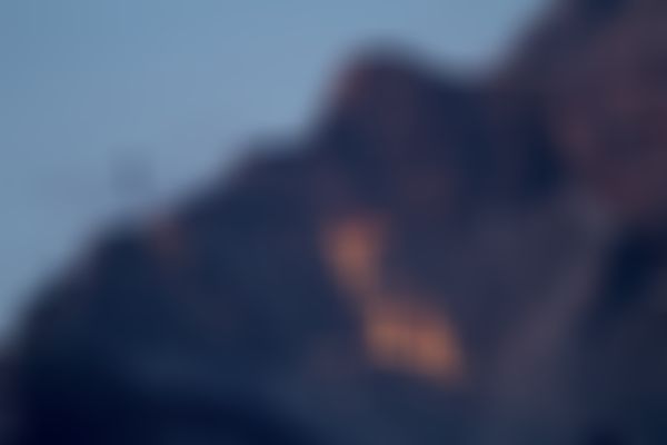 Ein Berg mit dem leuchtenden Umriss eines Steinbocks, beleuchtet von Lichtern, vor einem düsteren Himmel. Links ist eine Seilbahnlinie zu sehen.