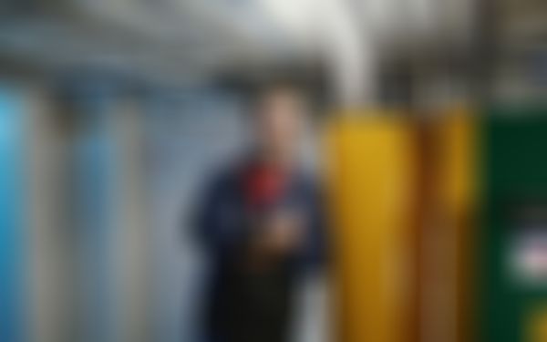 Junger Mann in dunkelblauer Arbeitskleidung der in einem Keller gegen eine Heizung lehnt.