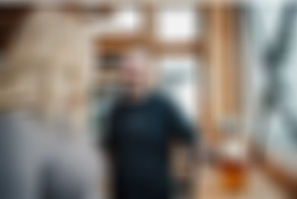 Lächelnder junger Kellner mit einem Bier in der linken Hand und einem Tablet in der rechten Hand. Davor eine junge blonde Frau mit Blick zum Kellner