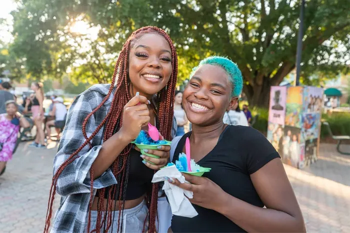 Two people smiling while holding colorful shaved ice in a sunny outdoor setting, with trees and people in the background.