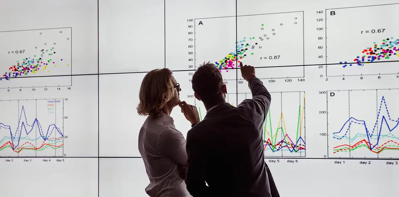 Two people analyze data on large screens displaying scatter plots and line graphs, discussing trends and correlations.