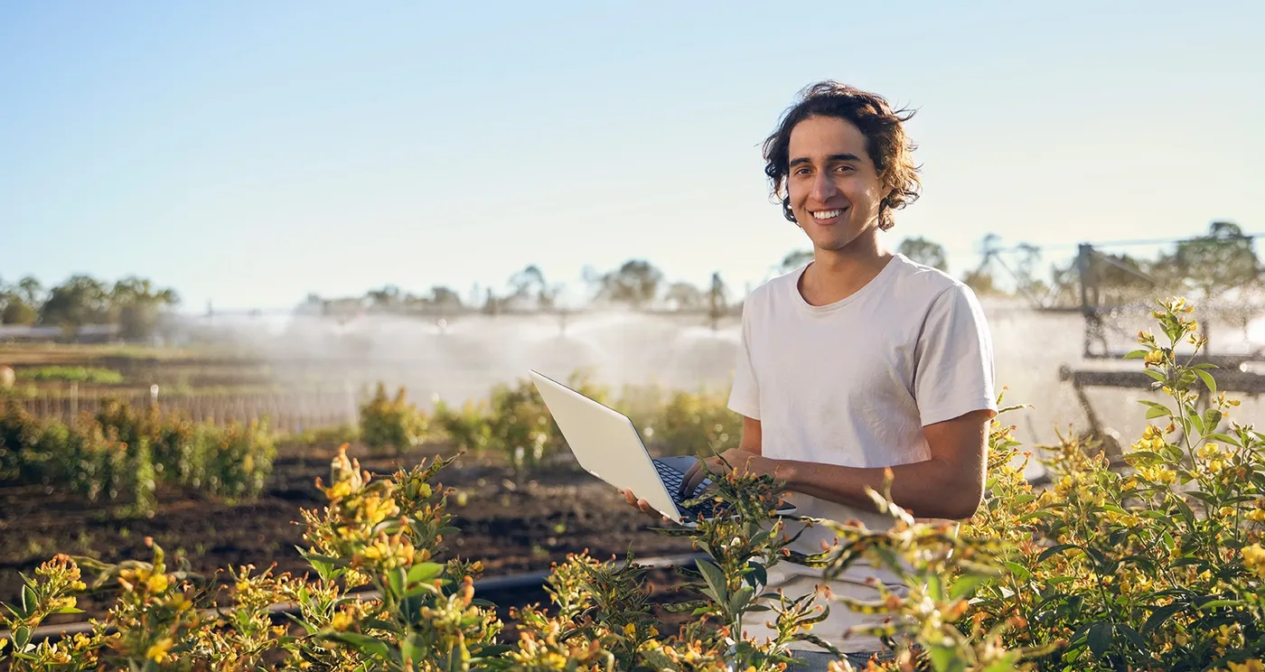 Person in a white shirt holding a laptop stands in a lush field with sprinklers in the background under a clear blue sky.