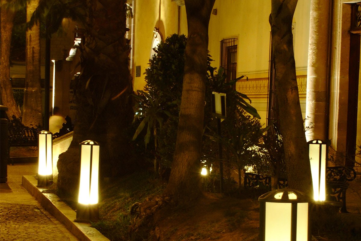 Paseo nocturno entre vegetación con balizas Simon JR1 alineadas, fachada colonial al fondo.