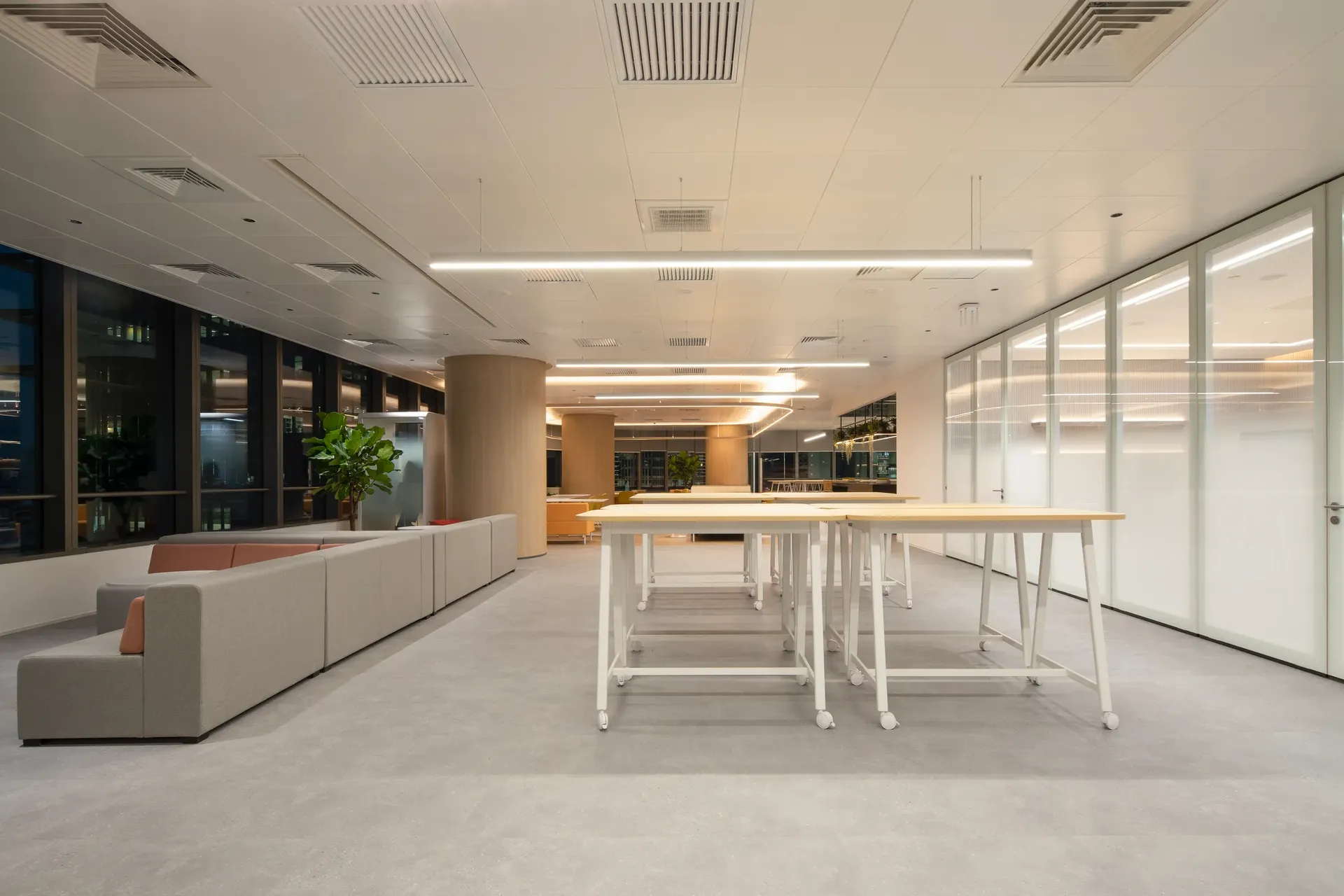 Oficina moderna en vista frontal con mesas de trabajo en primer plano y zona de sofás a la izquierda, mostrando un espacio abierto y luminoso de trabajo colaborativo.