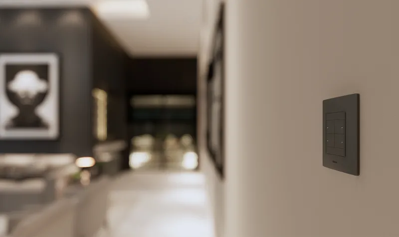 Lifestyle setting with a Simon 26 iO switch installed on a mustard-coloured wall, next to black decorative lanterns and a view to a window with natural light.