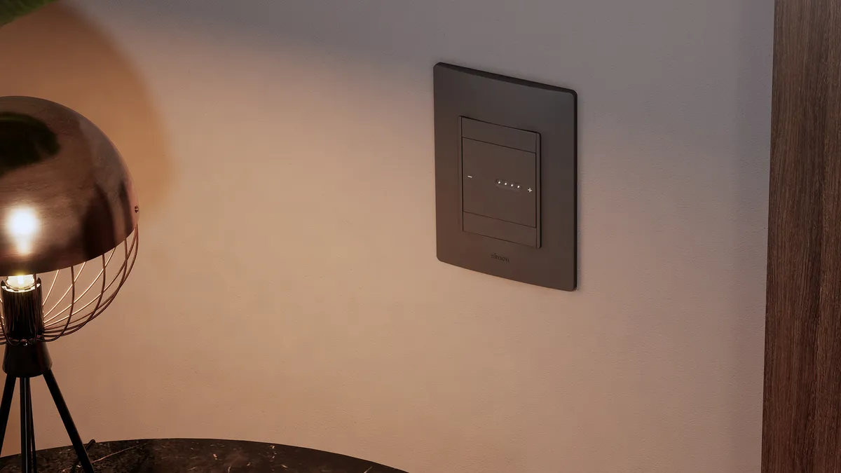 Simon 26 iO Dimmer — black dimmer switch installed on a wall with a copper lamp and a warm dimly-lit ambience.