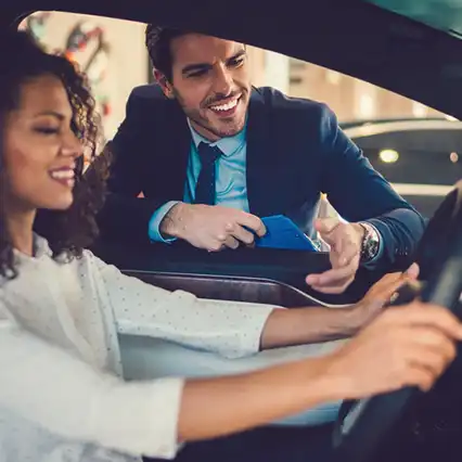 Een vrouw zit lachend in een auto, terwijl een man in pak door het raam naar binnen leunt, een blauwe map vasthoudt en met zijn hand gebaart.