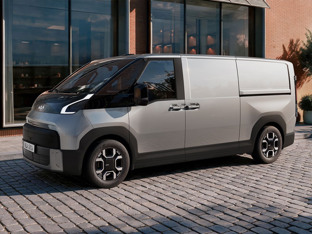 A modern, silver van parked on a cobblestone street in front of a building with large windows and greenery.