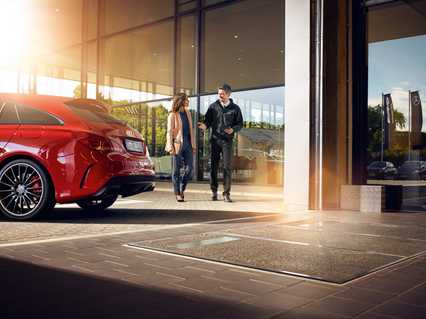 A salesman in casual attire stands outside a glass-walled car dealership, admiring a red sports car under a warm sunbeam.