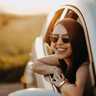 Woman smiling and wearing sunglasses, leaning out of a car window, with a scenic sunset in the background.