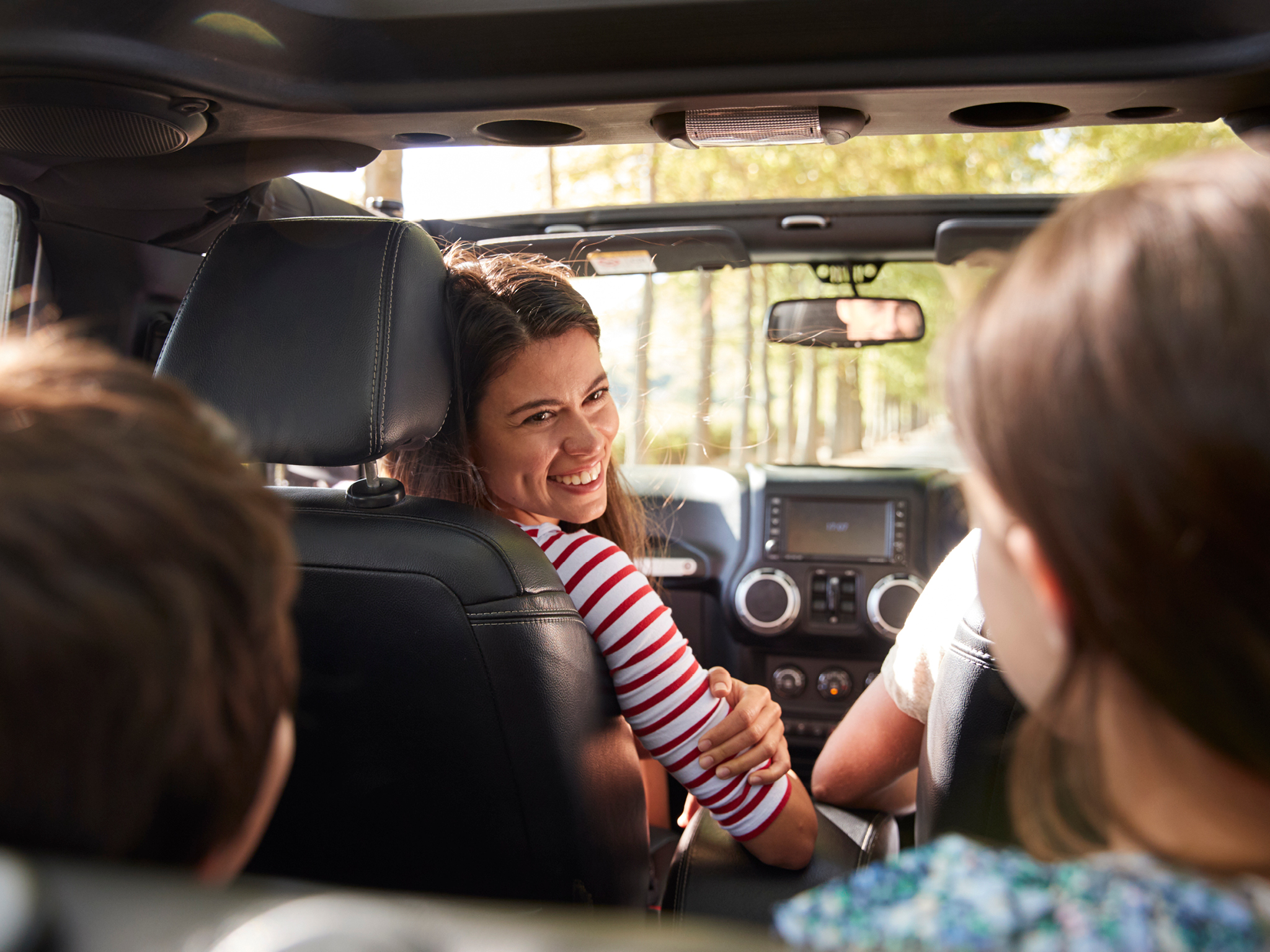 Woman smiling in a car with two children in the backseat, driving through a sunlit forest.