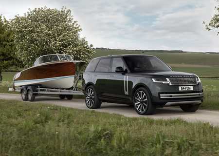 A Range Rover towing a wooden boat on a trailer along a country road, with green fields and trees in the background.
