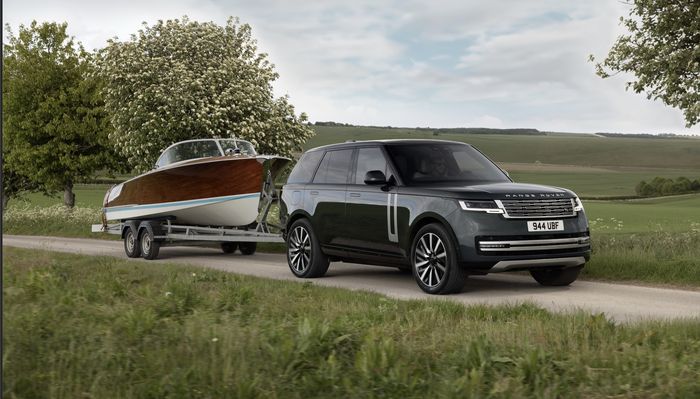 A Range Rover towing a wooden boat on a trailer along a country road, with green fields and trees in the background.