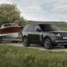 A Range Rover towing a wooden boat on a trailer along a country road, with green fields and trees in the background.
