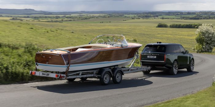 An electric Range Rover towing a wooden speedboat on a trailer through a scenic countryside road with green fields in the background.