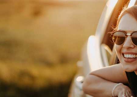 Woman with sunglasses smiling, leaning out of a car window, enjoying a sunny day with a blurred green field in the background.