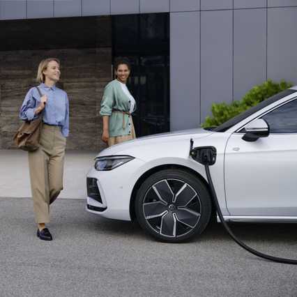 Two women walking past a white electric car being charged, parked in front of a modern building.