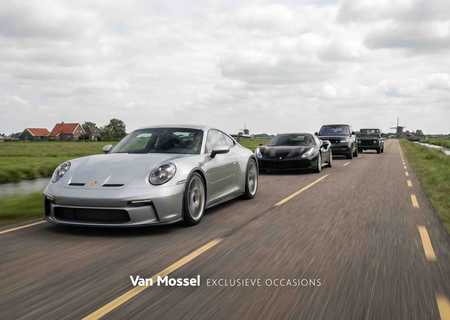 Four luxury cars drive in a line on a rural road, with green fields and a cloudy sky in the background.