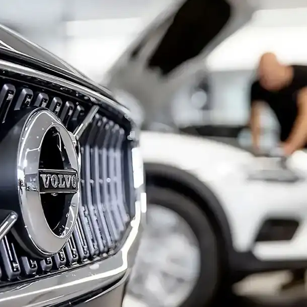 Close-up van de grille van een Volvo-auto met scherp logo, terwijl iemand een andere auto met een open motorkap op de onscherpe achtergrond inspecteert.