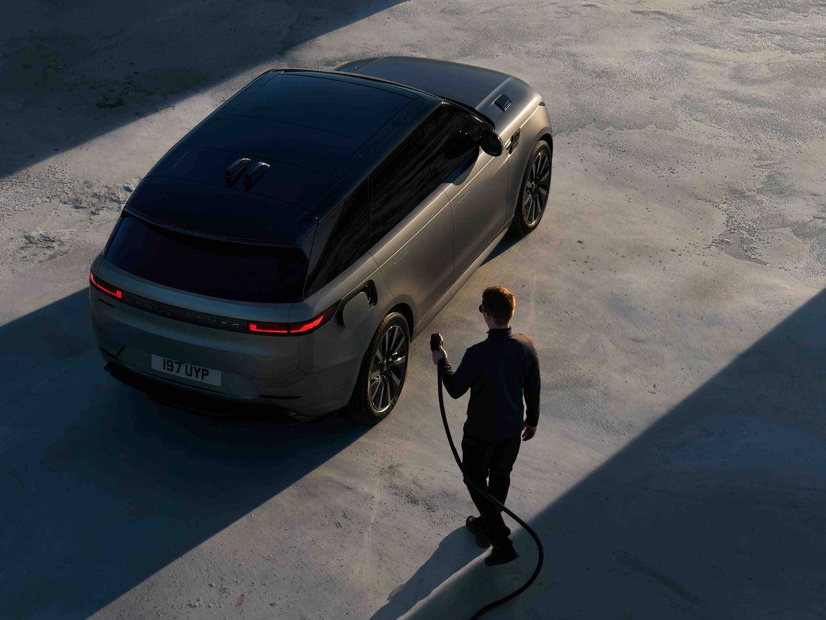A man approaches a sleek, dark SUV with an electric charging cable against a shadowed concrete surface.
