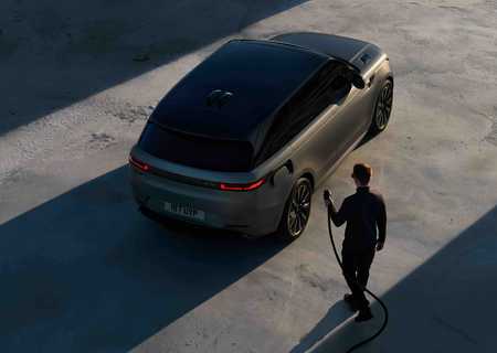 A man approaches a sleek, dark SUV with an electric charging cable against a shadowed concrete surface.