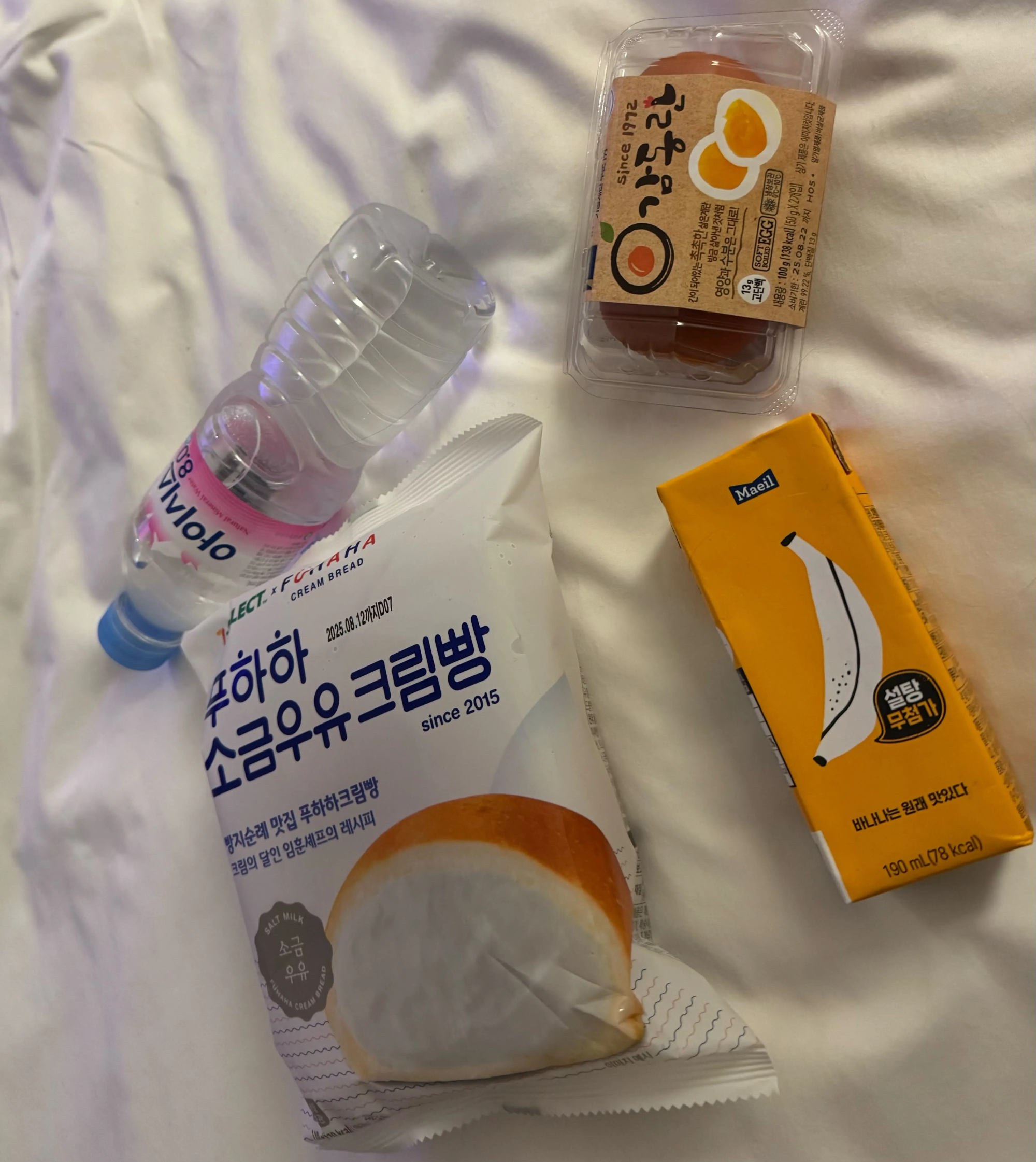 A bottle of water, a pack of marinated boiled eggs, a giant cream bun and a box of banana milk on a hotel bed.