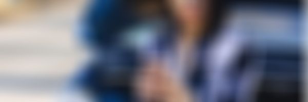 Junge Frau sitzt vor offener Motorhaube und schaut ins Handy.