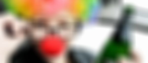 Frau als Clown verkleidet mit bunter Perücke, roter Nase und Brille mit einer Prosecco-Flasche mit null Promille in der Hand.