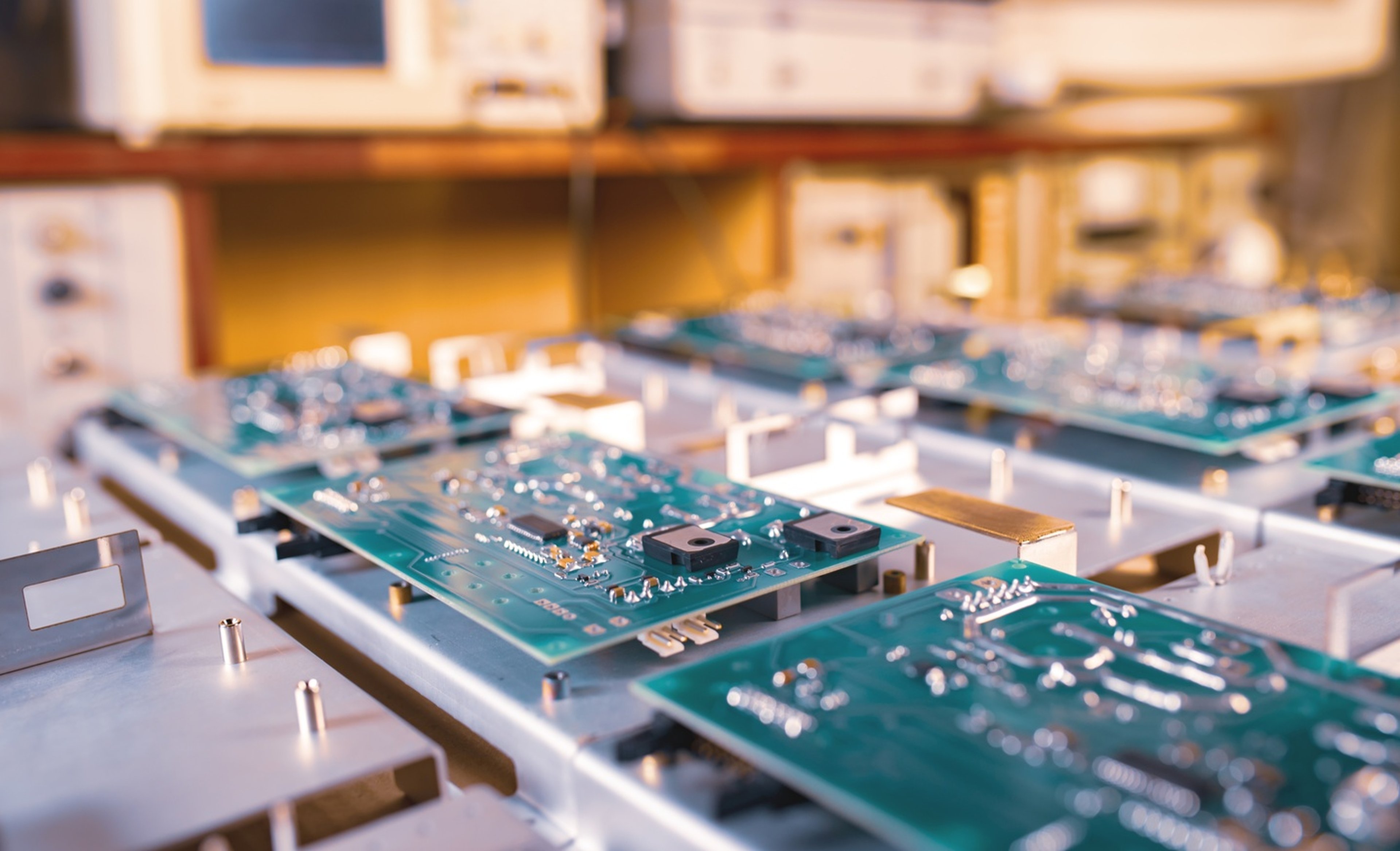 Close-up view of multiple green circuit boards moving along a production line in a factory, with blurred electronic equipment in the background, illustrating precision assembly and high-volume electronics manufacturing.
