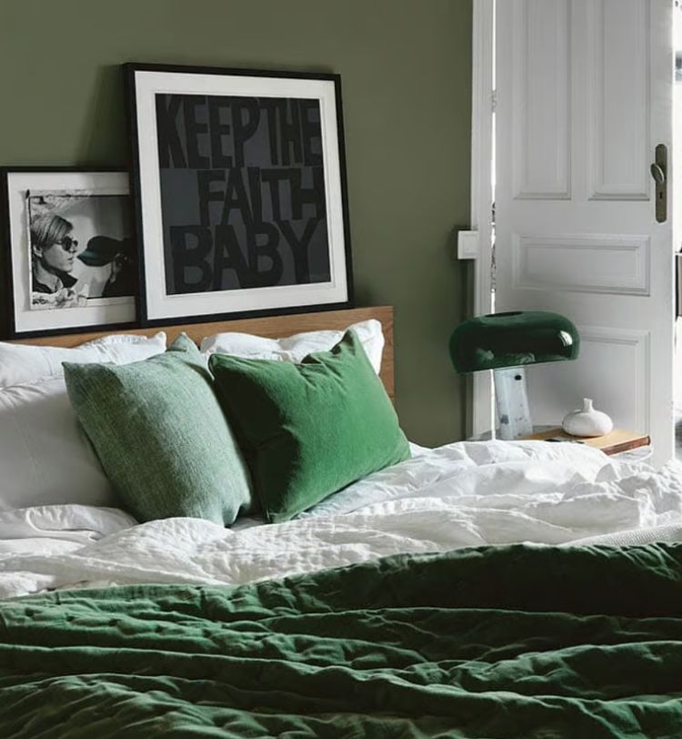 Cozy bedroom with green bedding, two green pillows, and framed art on a wooden headboard against an olive wall. White door slightly ajar.