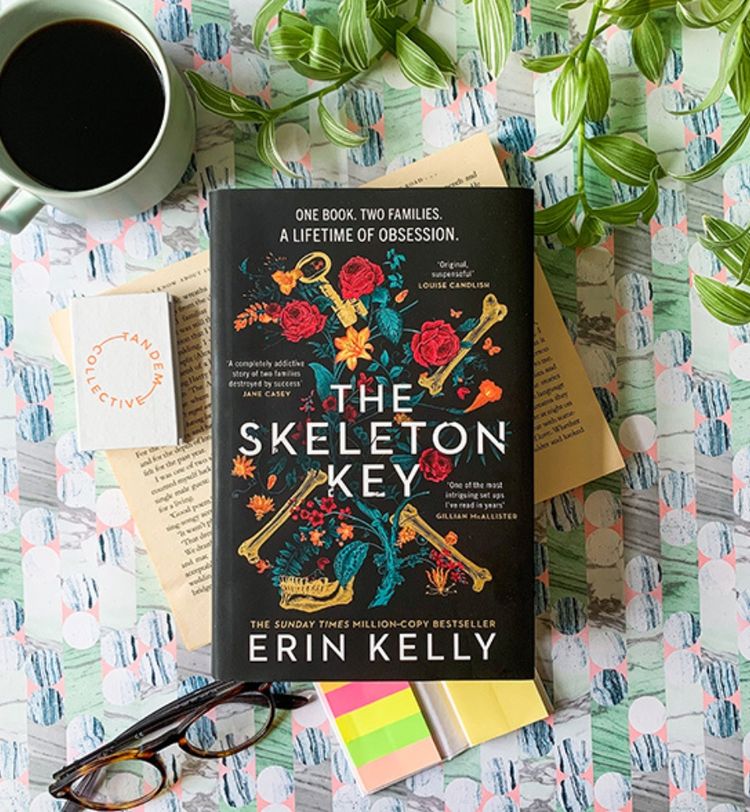 A book titled "The Skeleton Key" by Erin Kelly on a table with a coffee cup, glasses, and a plant. The cover features floral and key designs.