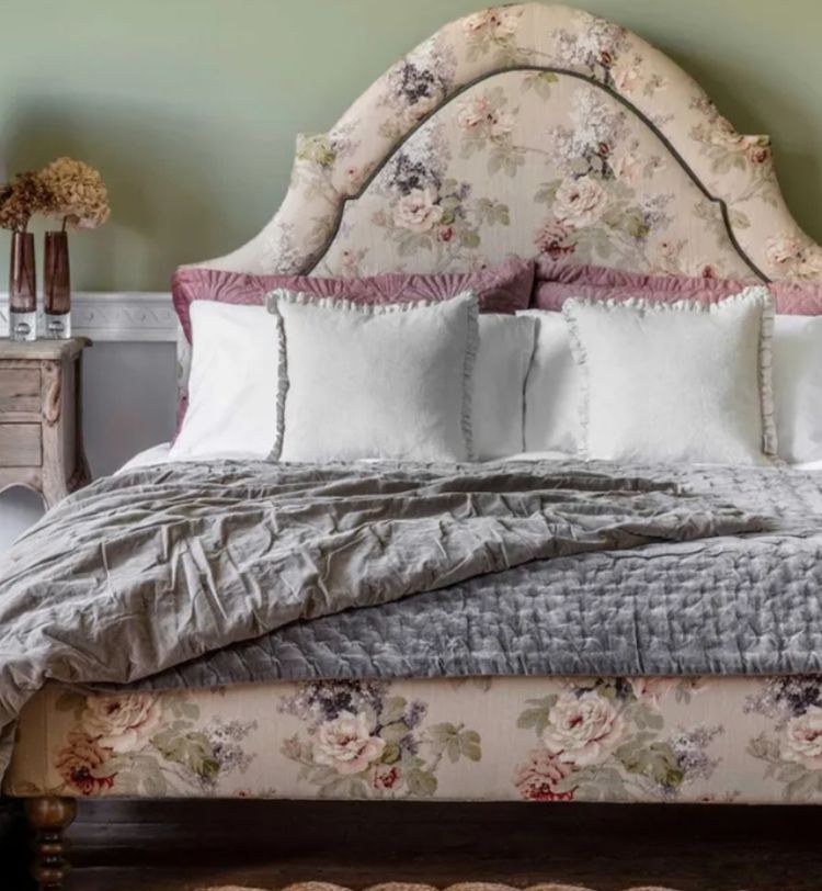 Floral-patterned bed with plush pillows and a gray quilt, flanked by a wooden nightstand with a lamp and dried flowers, against a green wall.