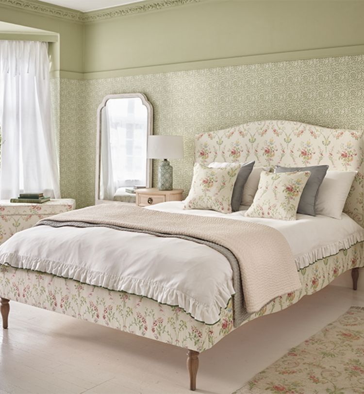 Elegant bedroom with floral-patterned bed, matching pillows, and light green wallpaper. A mirror, lamp, and window with sheer curtains complete the decor.