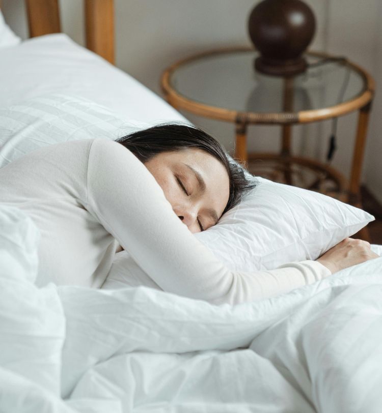 A person sleeping peacefully on a white bed, with a wooden nightstand and lamp in the background.