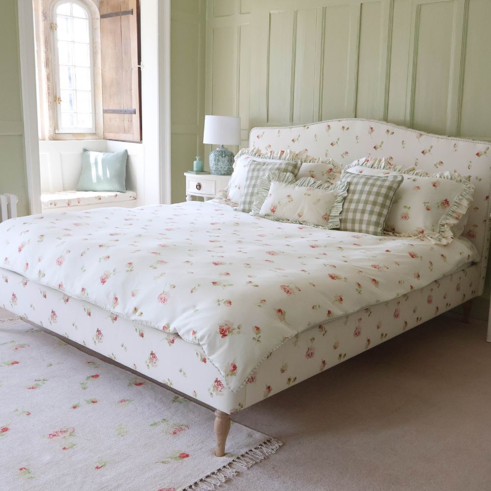 Cozy bedroom with a floral-patterned bed, matching pillows, and a soft rug. Light green walls and a window with a cushioned seat.