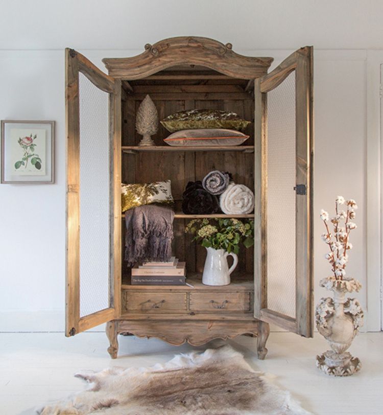 Open wooden cabinet with shelves holding folded blankets, pillows, and a vase. A framed flower print and decorative vase are nearby.