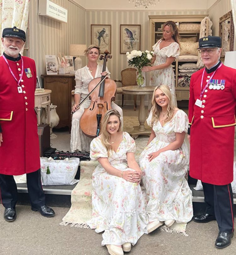 Two men in red uniforms stand with four women in floral dresses, one playing a cello, in a vintage-style room with floral decor.