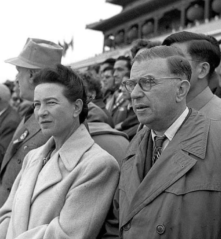 A black-and-white photo of a man and woman in coats standing among a crowd, appearing to watch an event.