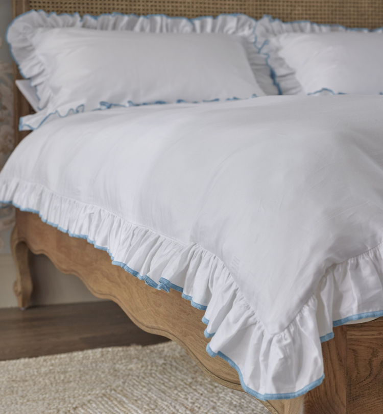 White bed with ruffled edges and light blue trim, wooden frame, and matching pillows on a textured rug in a cozy bedroom setting.
