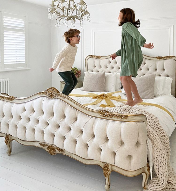 Two children joyfully jumping on an elegant, tufted bed in a bright room with a chandelier and white walls.