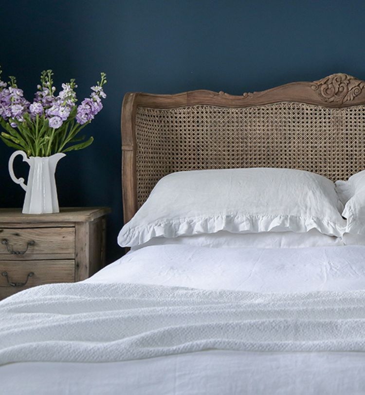Cozy bedroom with a wooden headboard, white bedding, and a bedside table holding a white vase of purple flowers against a dark blue wall.