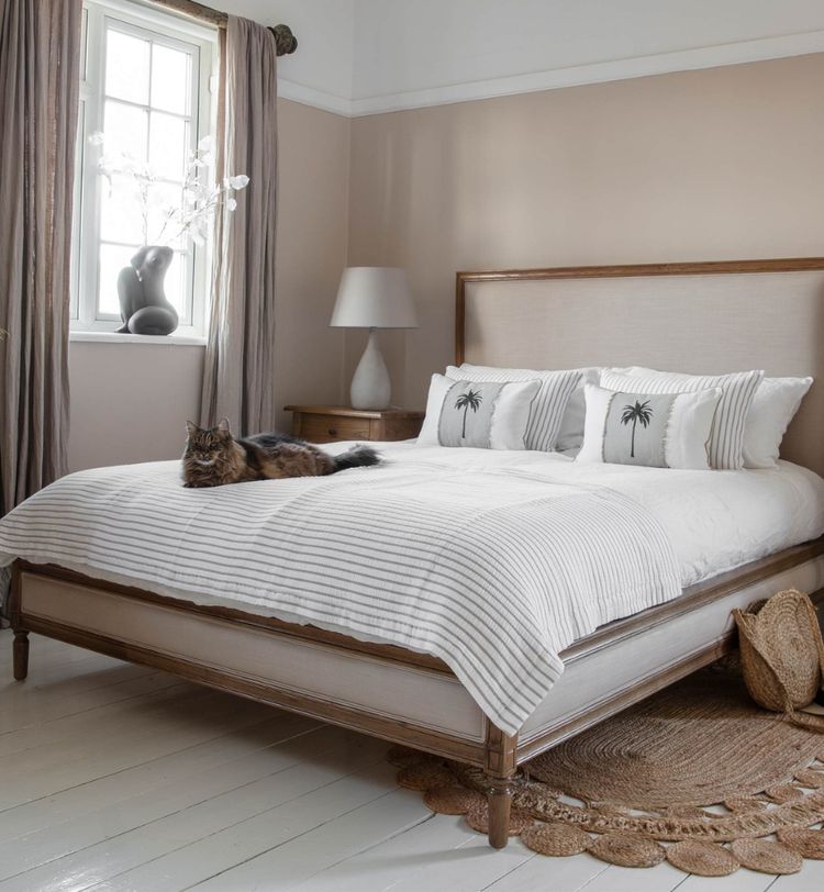 Cozy bedroom with a large bed, white and striped bedding, decorative pillows, a cat lounging, a lamp, and a window with curtains.